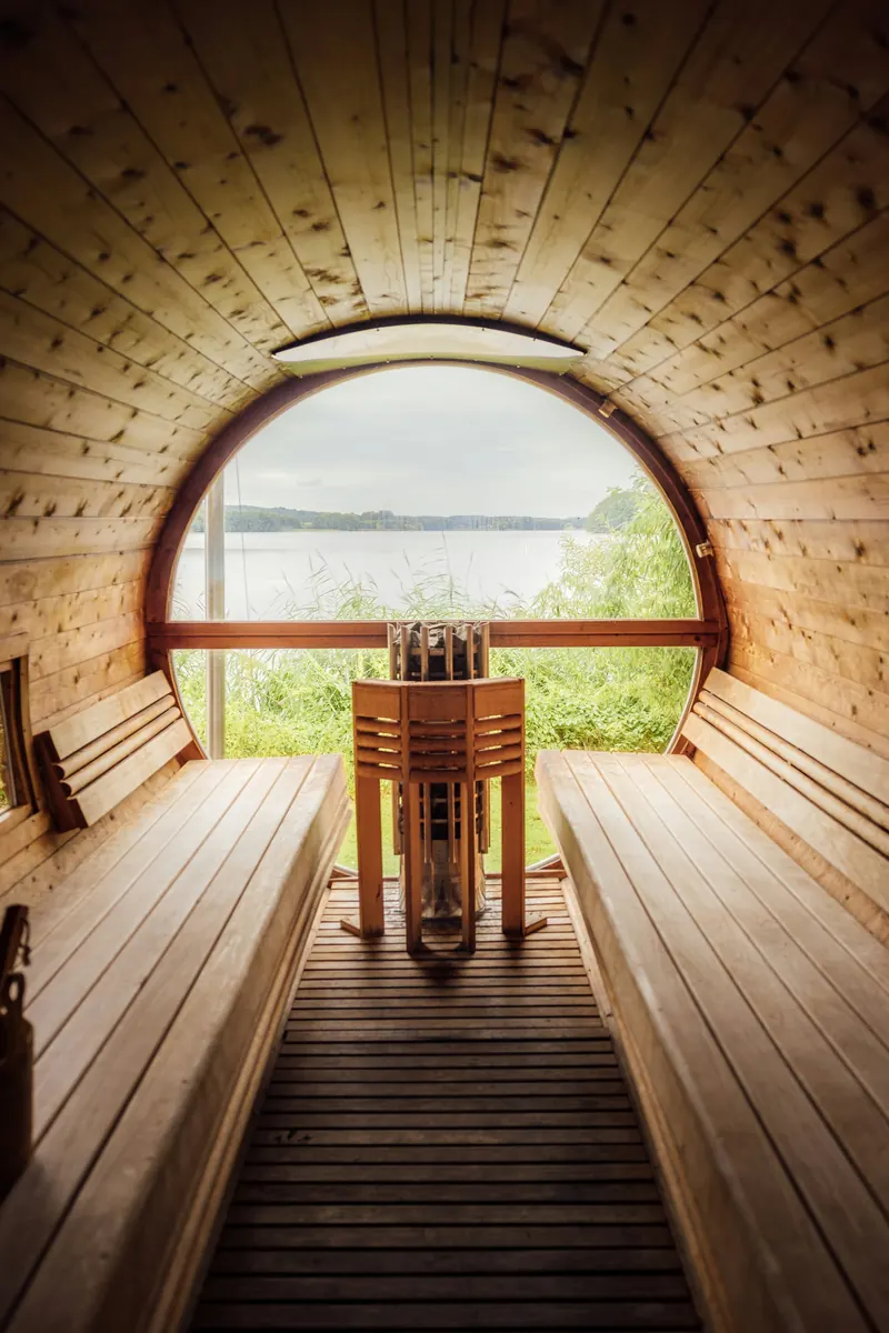 Inside a barrel sauna: two benches, a heater with a wooden guard and a round window onto a lake