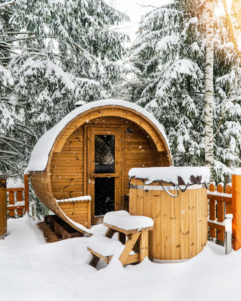 A barrel sauna and a wooden hot tub covered in snow among spruce trees